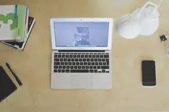 Ahşap masada açık dizüstü bilgisayar, telefon, defter ve kitaplar, yukarıdan