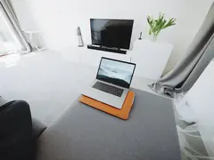 Beyaz oturma odasında televizyon ünitesi, soundbar ve hoparlör; önde koltukta dizüstü bilgisayar